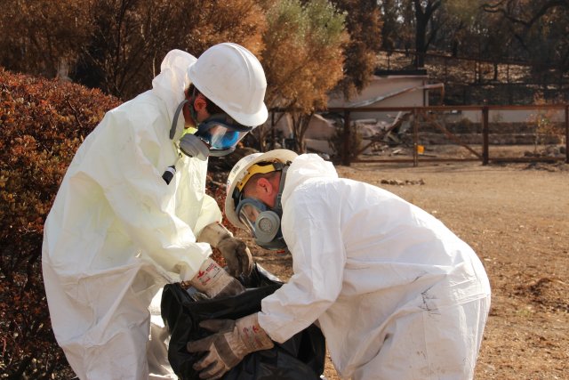 Two workers in hazardous material gear placing waste into garbage bags.