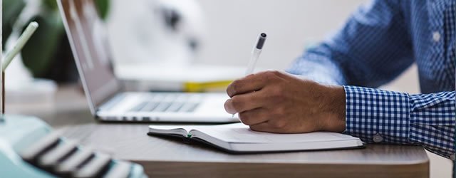 Man writing in a notebook with a laptop in the background.