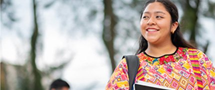 A young person smiling, wearing a backpack and holding a book