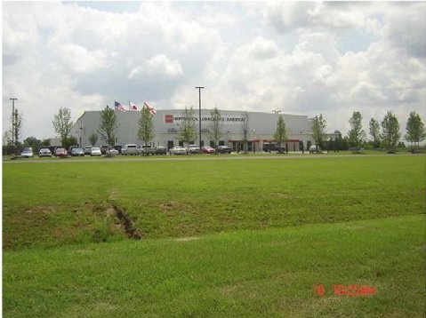 A grassy field with a parking lot, building, and cloudy sky in the background.