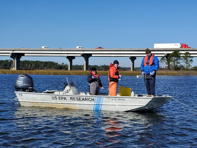 Three people with lifejackets on a boat, which says "US EPA Research,' in a body of water.