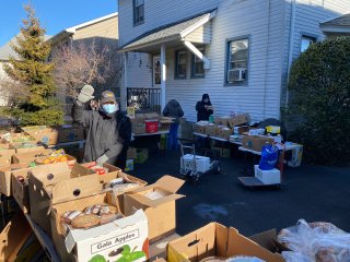 Image of a gentleman waving at the camera outside of a house with boxes of food available. 