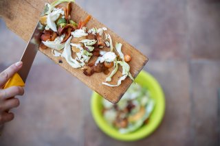 Image of a person using a knife to scrape vegetable scraps off a cutting board into a green bucket.