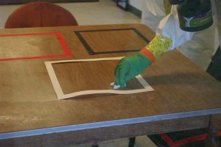 Responder wearing a green glove uses a wipe to sample a wooden table inside a white square. There is an additional red square and black square outline next to it.