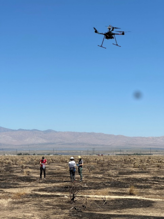 Three people stand in an arid landscape with a drone flying overhead.