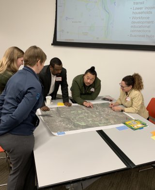 Image shows a group of people huddled around a table with a map on it, in a community workshop.