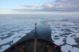 Bow of large arctic vessel surrounded by chunks of sea ice against a deep blue ocean, as far as the eye can see.