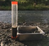 A stream with a rocky shore. There is a metal container with silt sitting on the shore with next to a test beakers with a  water and sediment sample.