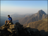 Sam Suzuki sits on a mountain top in South Africa with a vista of mountains behind him