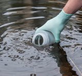 Gloved hand holding a bottle and collecting a sample from a body of water 